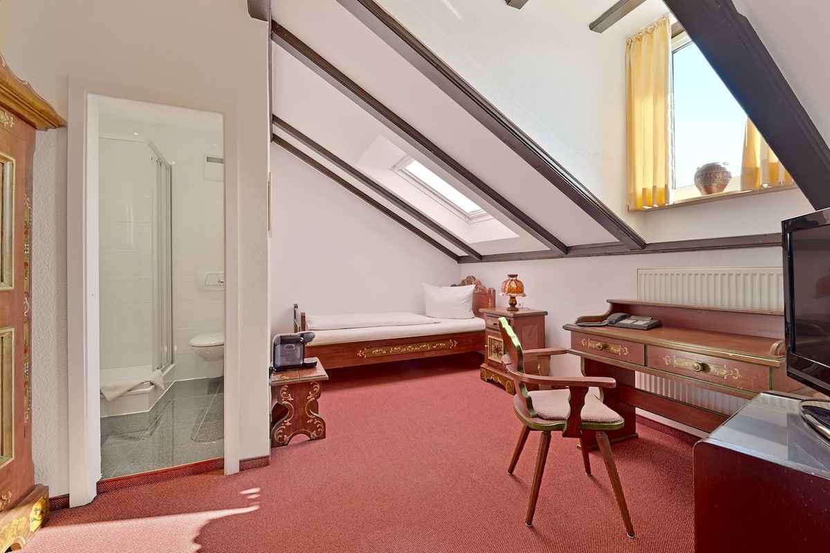 A bedroom with a sloped ceiling, a bed, a desk, a chair, a television, and a window with curtains.