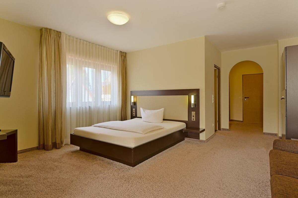 A hotel room with a large bed, a desk, a chair, and a door leading to another room.