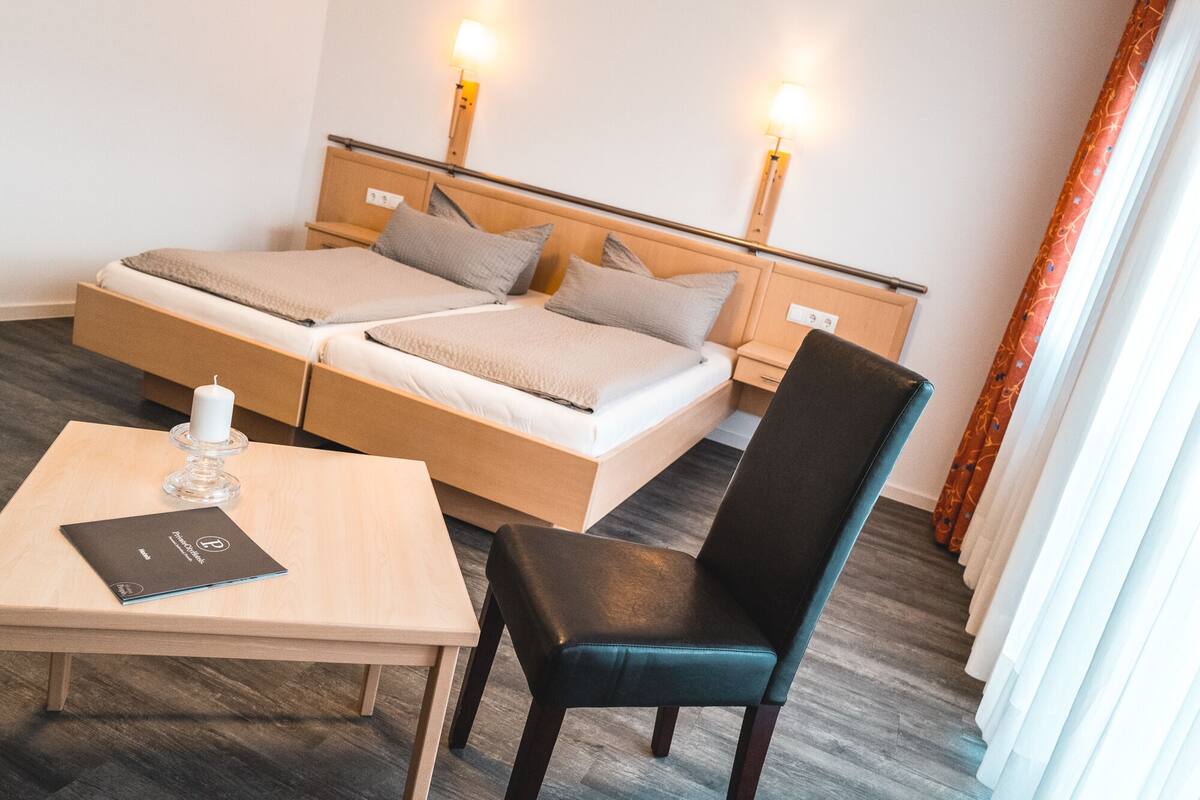 A double bed with a wooden headboard, a bedside table with a lamp, a chair, and a small table with a book.