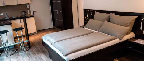 A modern bedroom with a large bed, a small kitchenette, and a separate bathroom area.