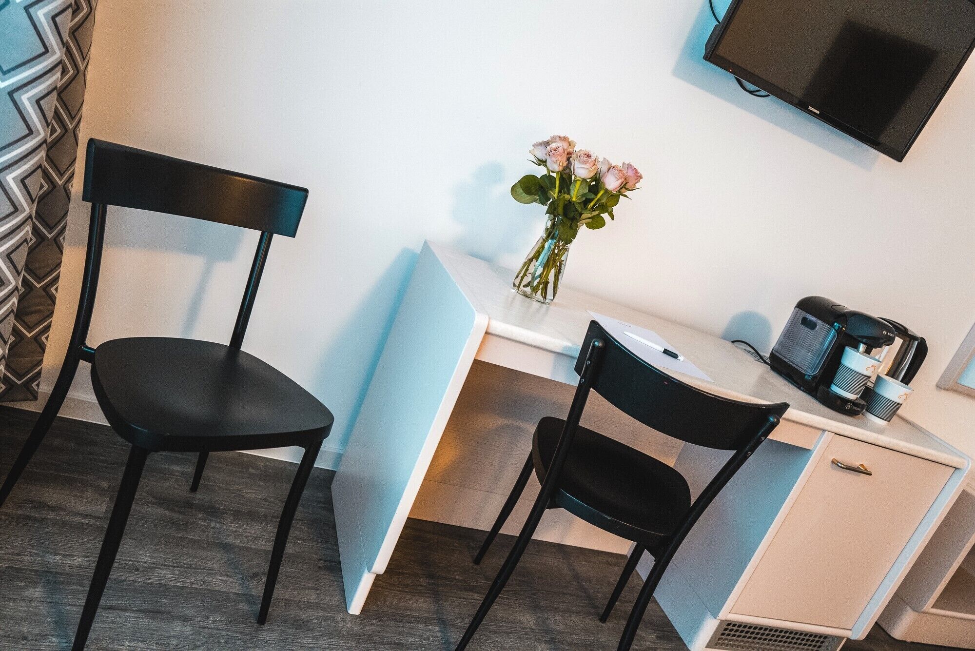 A hotel room with two black chairs, a small desk with a coffee maker, a TV mounted on the wall, and a vase with flowers on the desk.