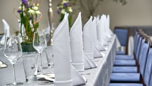 Ein Esstisch ist gedeckt mit weißen Servietten, Weingläsern und Besteck, im Hintergrund sind Stühle und ein Blumenstrauß zu sehen.
