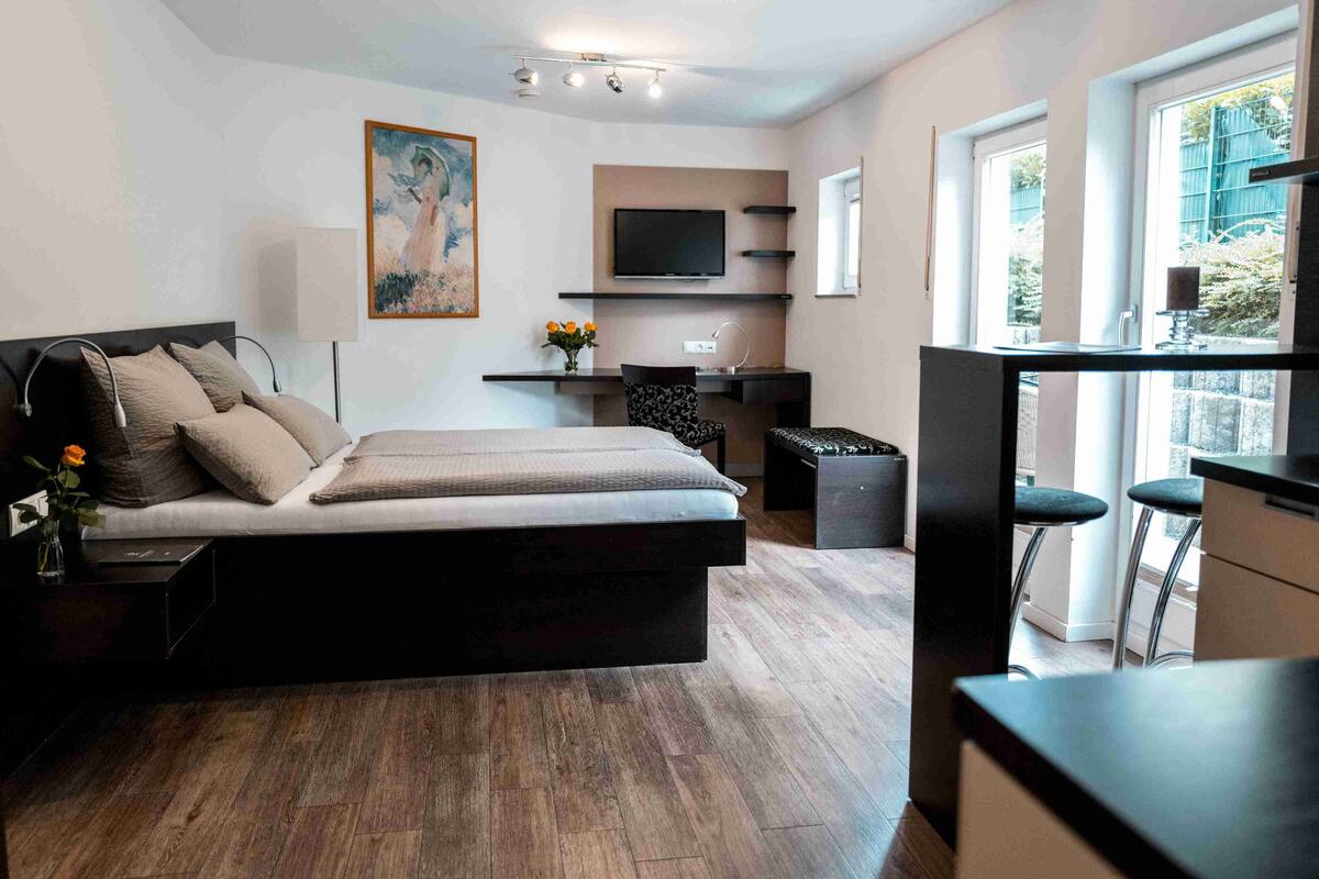 A modern bedroom with a large bed, a desk, a TV, and a window with a view of greenery.