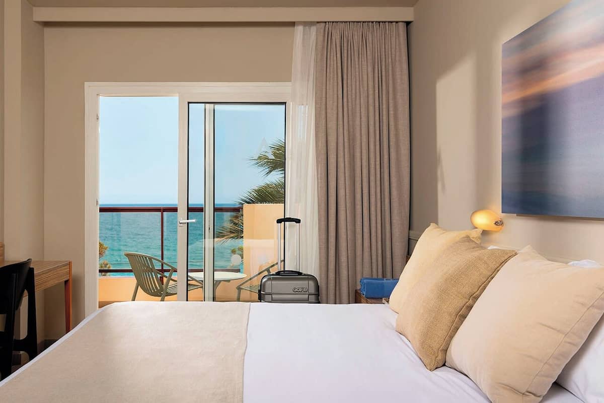A hotel room with a bed, a suitcase, a nightstand, a lamp, a view of the ocean, and a balcony with a chair and table.