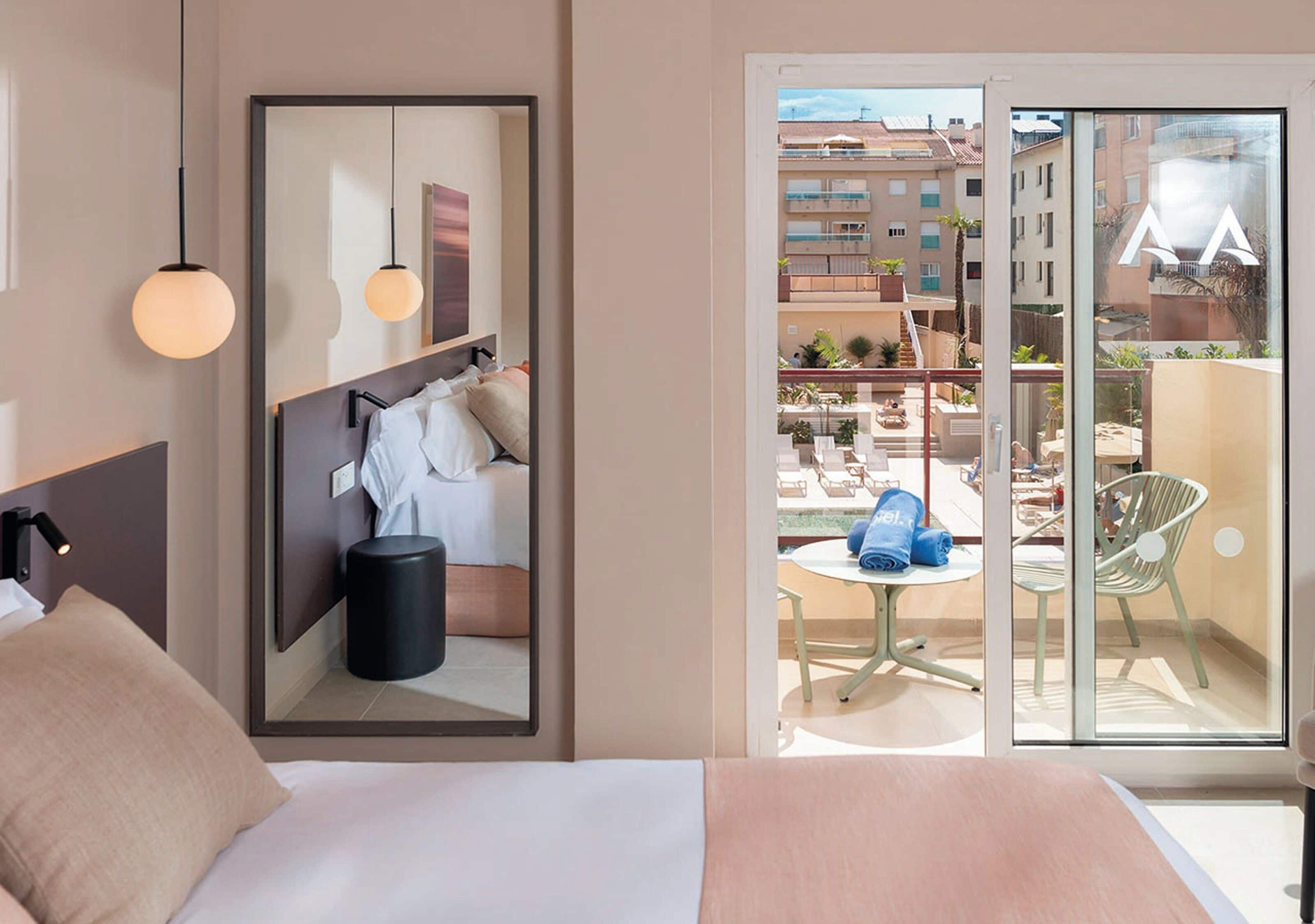 Een hotelkamer met een bed, een spiegel, een klein tafeltje met een stoel, en een balkon met uitzicht op de stad.