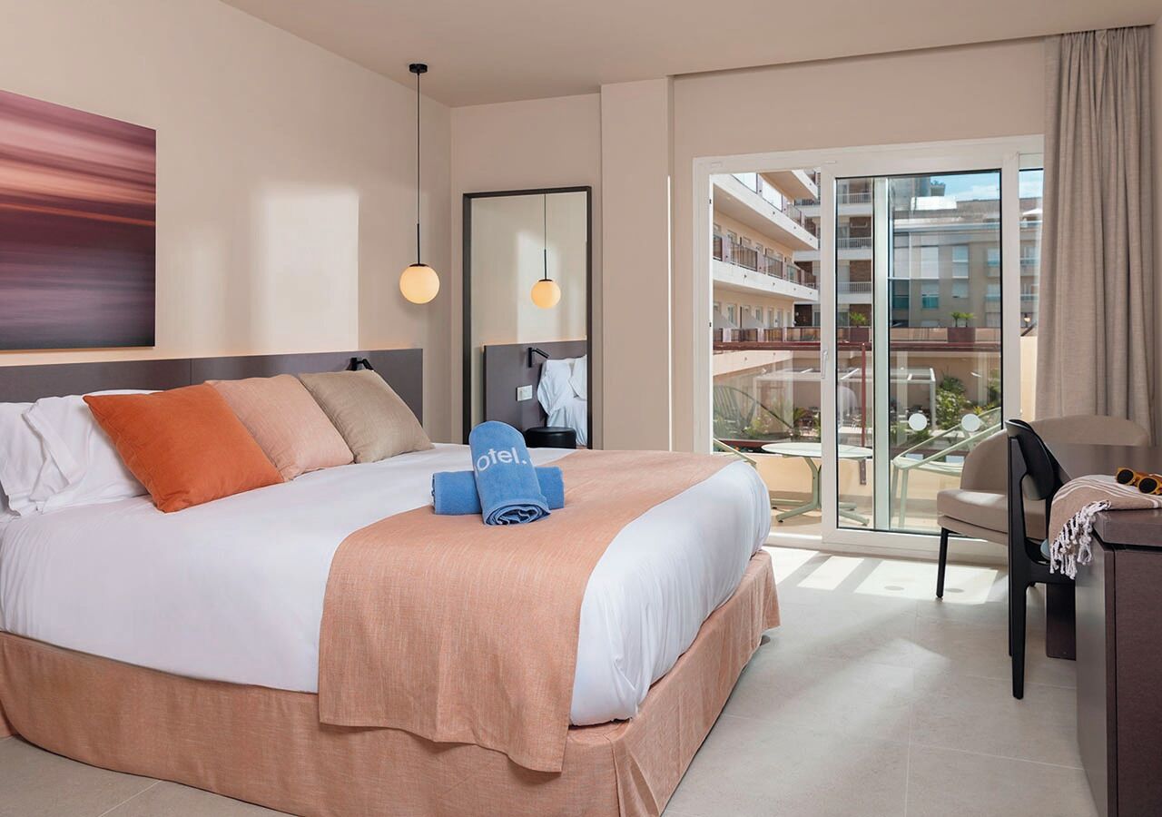A hotel room with a bed, a desk, a chair, a large window, and a balcony.
