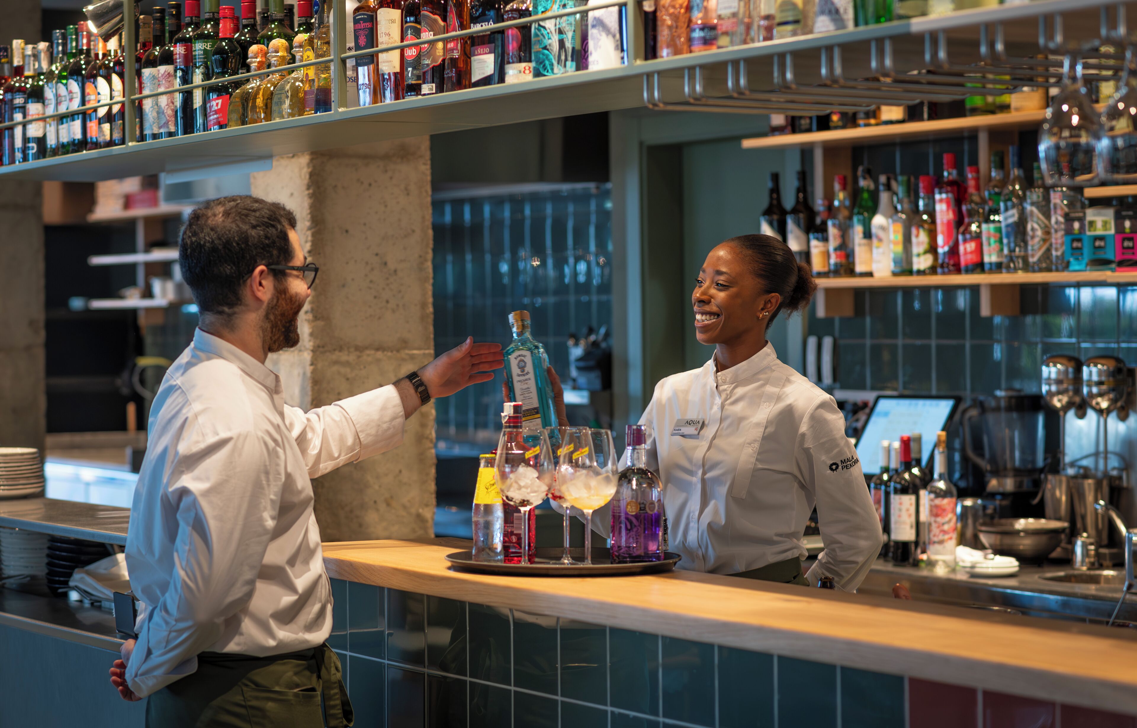 Zona de bar con estantes llenos de diferentes botellas, un bartender preparando bebidas y un cliente interactuando con él.