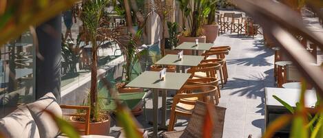 An outdoor café with tables and chairs, surrounded by potted plants and a tiled floor.