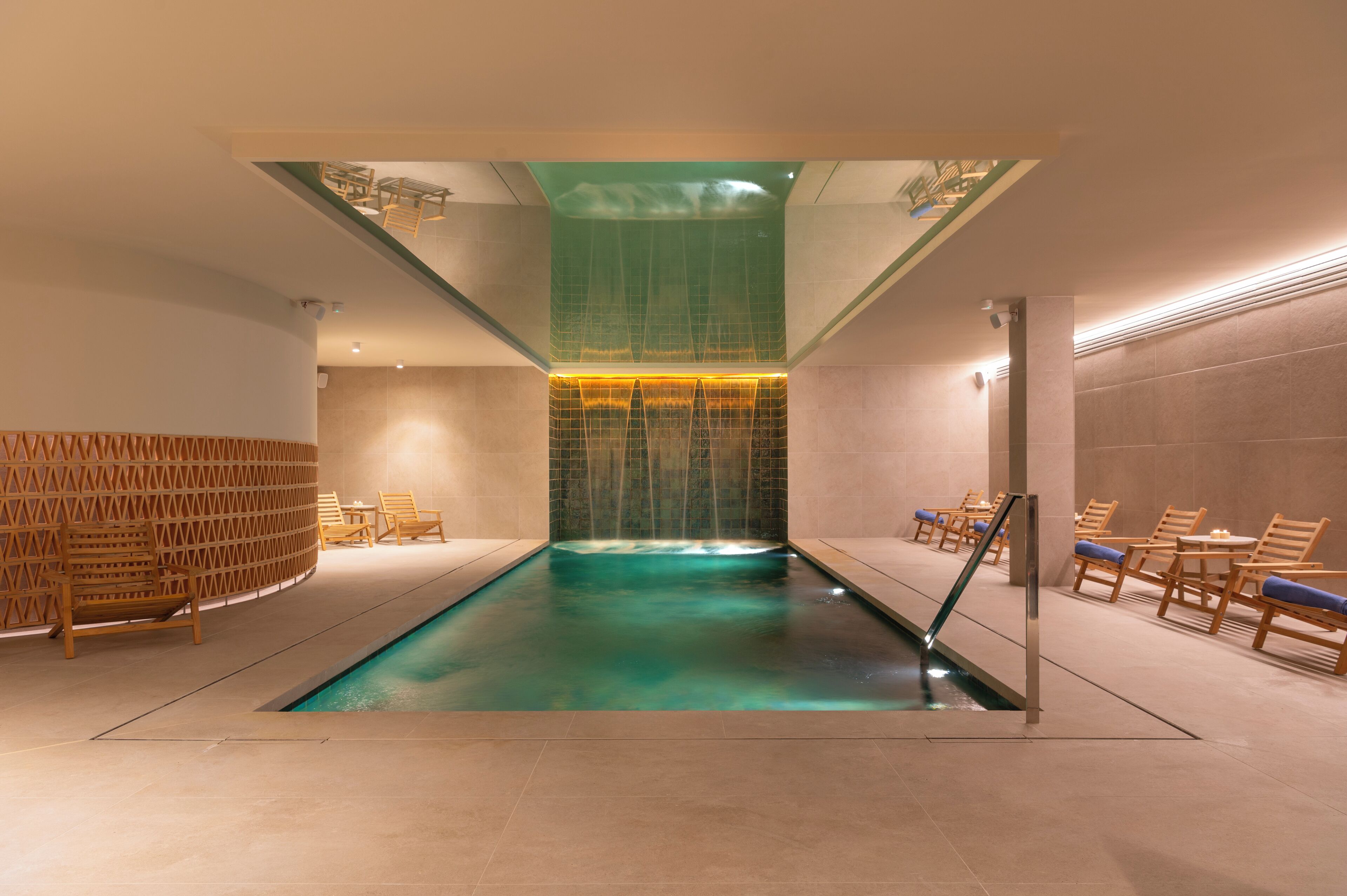 En moderne swimmingpool indendørs med klart vand, omgivet af træstole og en glasafskærmning.