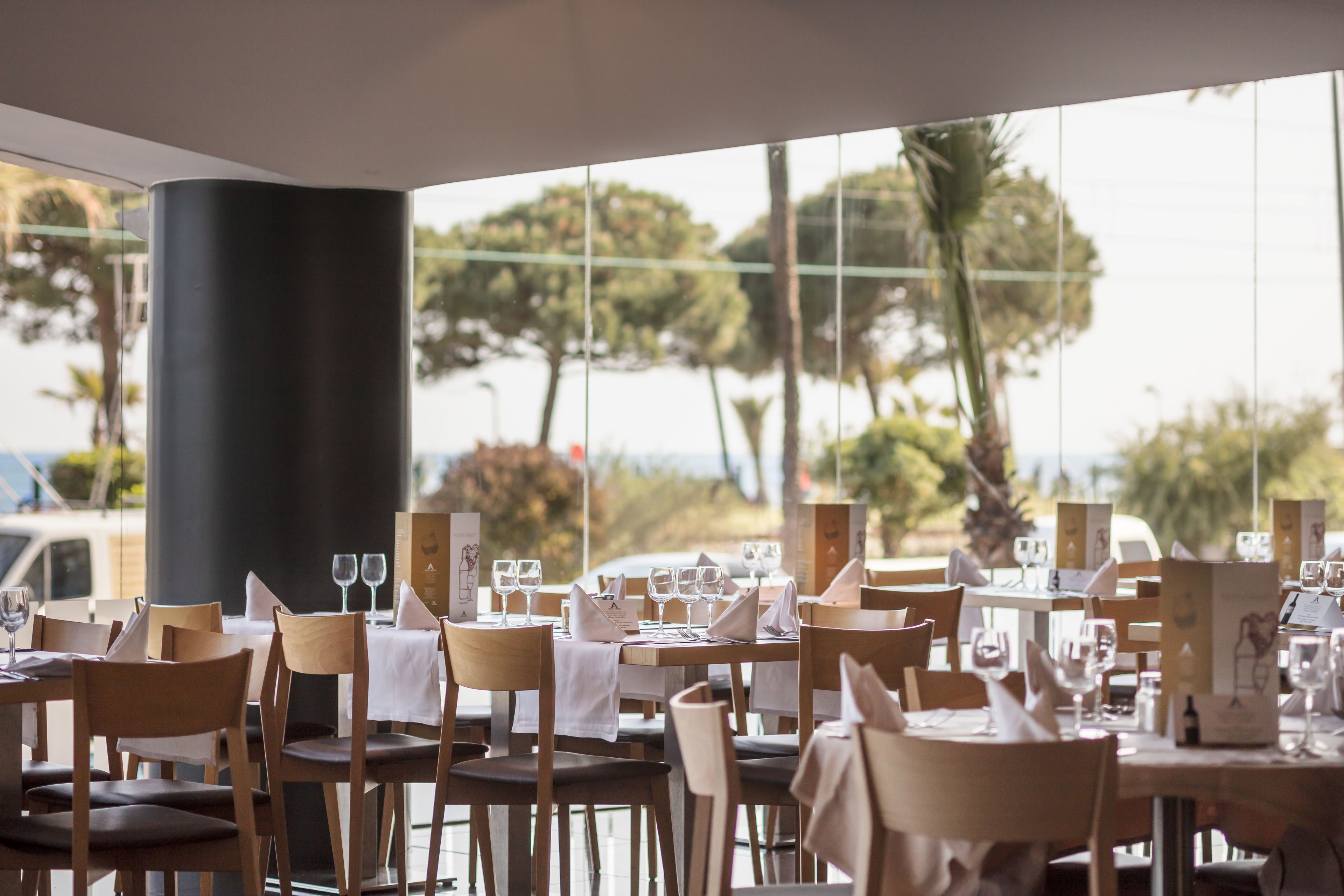 A restaurant with tables set for dining, featuring a view of palm trees and a body of water through large windows.