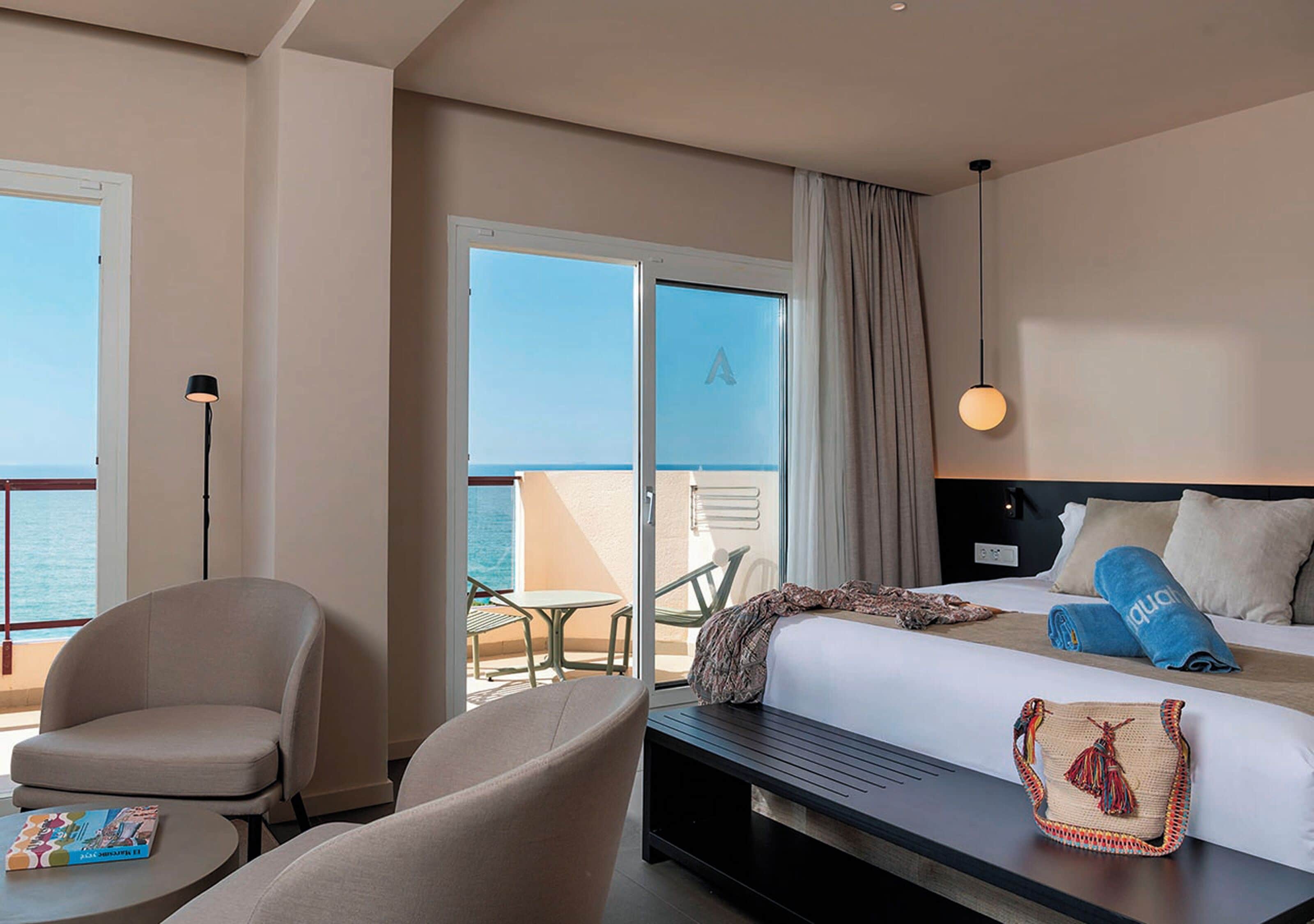 A hotel room with a bed, a desk, a chair, and a view of the sea.
