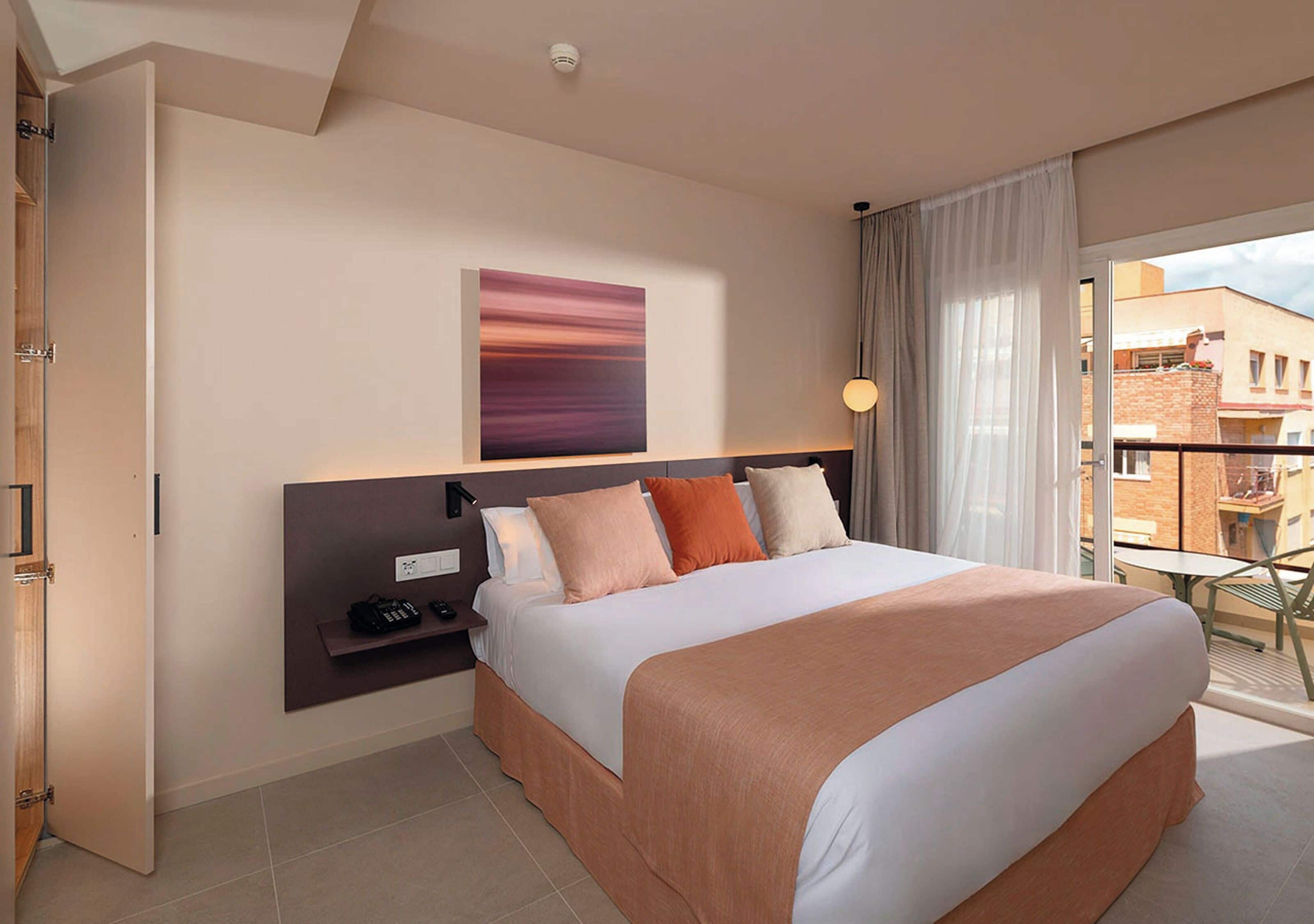 A modern hotel room with a large bed, a bedside table, a wall-mounted TV, and a view of the outside balcony.
