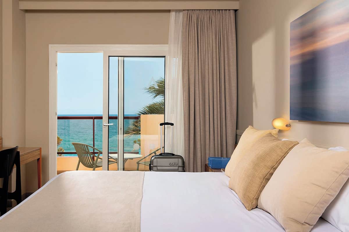 Une chambre d’hôtel avec un lit, une valise, une chaise et une vue sur l’océan, visible à travers une porte coulissante.