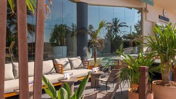 A modern outdoor seating area with a sofa, chairs, and tables, surrounded by potted plants and palm trees.