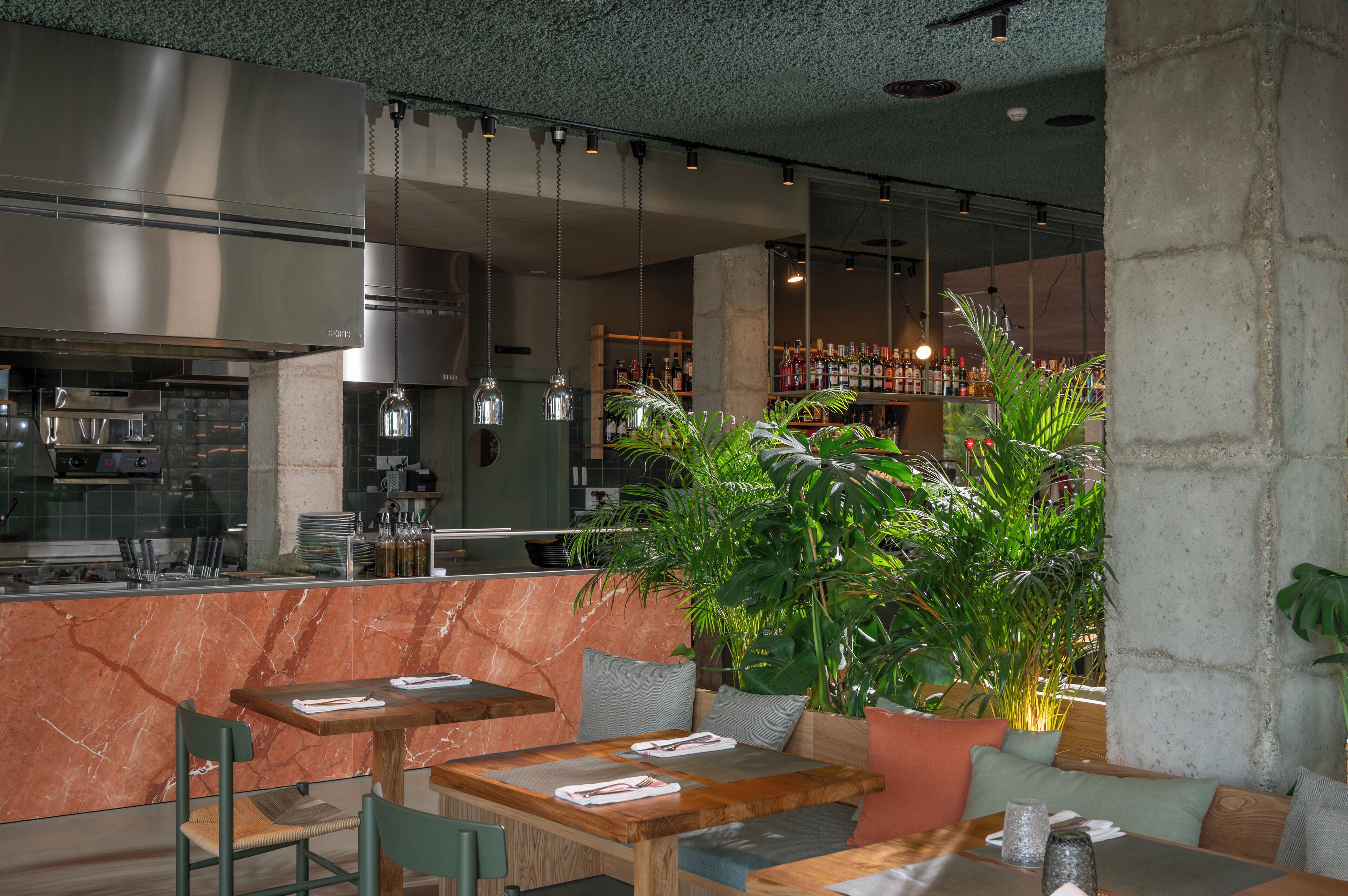 Un restaurante con mostradores de mármol, mesas de madera y sillas verdes. Se observan luces colgantes y una zona de bar al fondo.