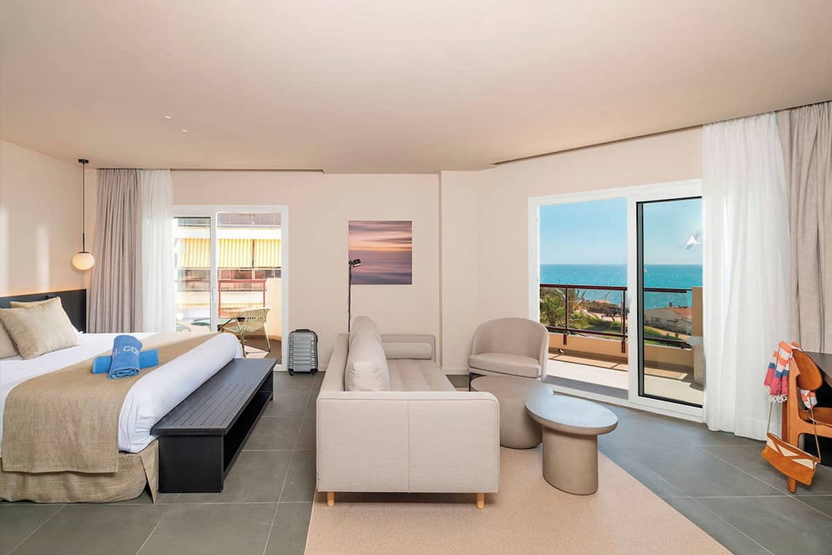 Een moderne hotelkamer met een bed, bank, fauteuil en een kleintje tafeltje. Er is een balkon met uitzicht op zee.