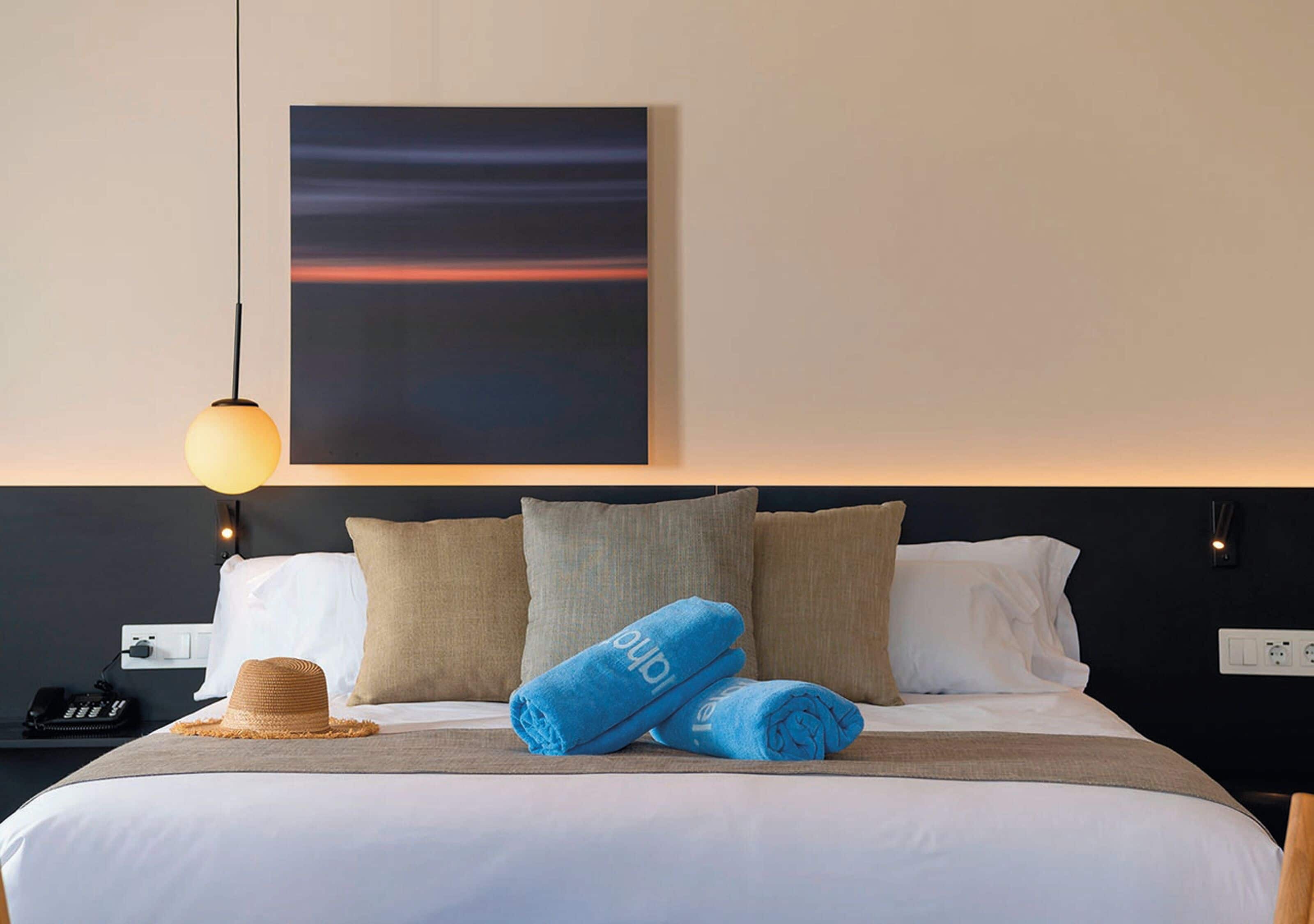 A neatly made bed with blue towels and a straw hat, a painting above it, and a hanging light fixture.