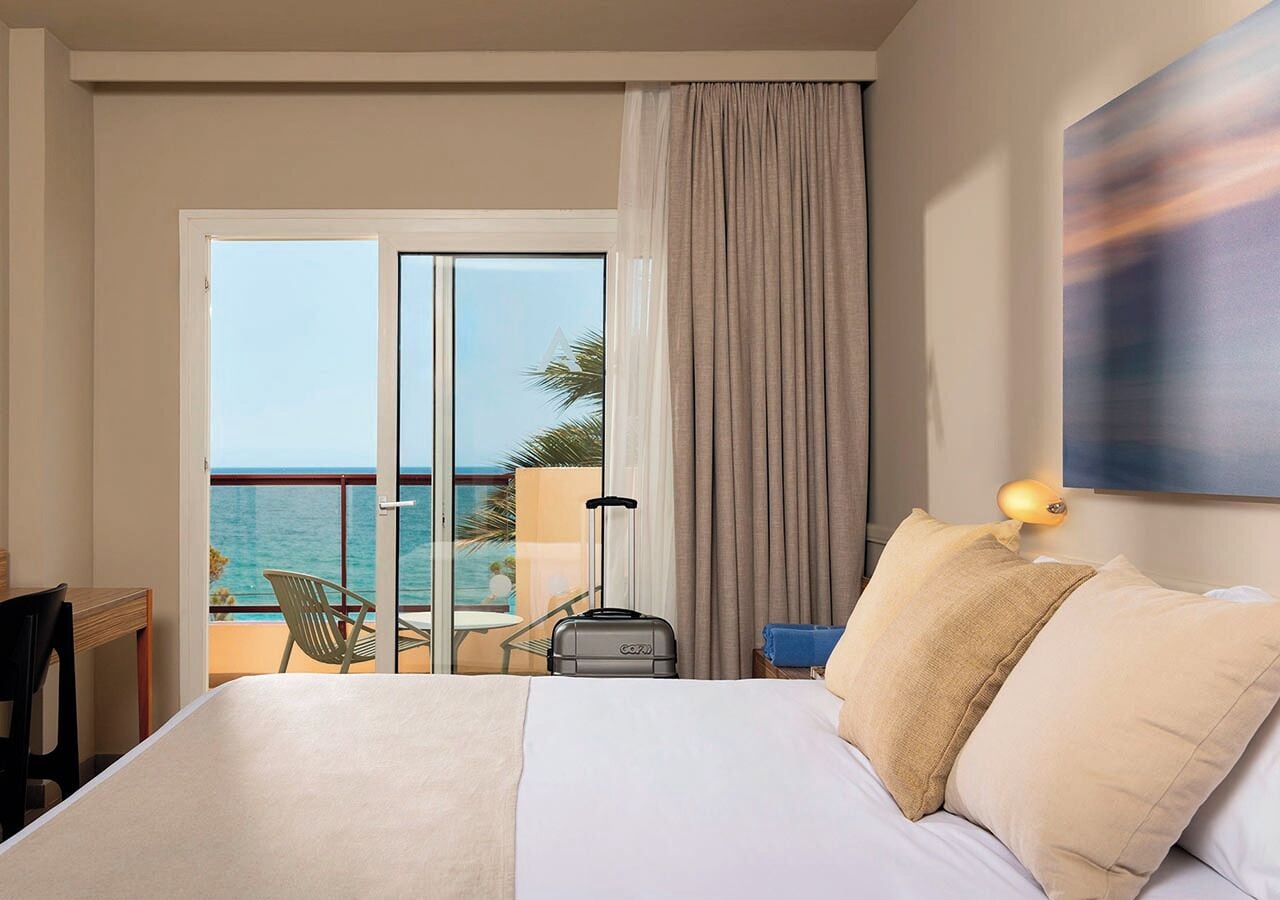 A hotel room with a bed, a suitcase, a nightstand, a lamp, a view of the ocean, and a balcony with a chair and table.