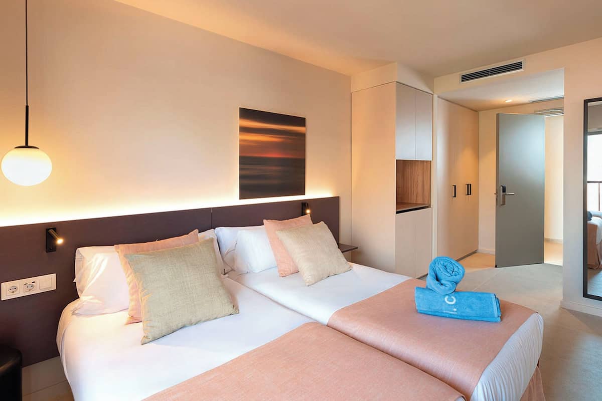 Une chambre d’hôtel avec deux lits, une tête de lit, une œuvre d’art accrochée au mur, une serviette sur le lit et une porte donnant sur une autre pièce.