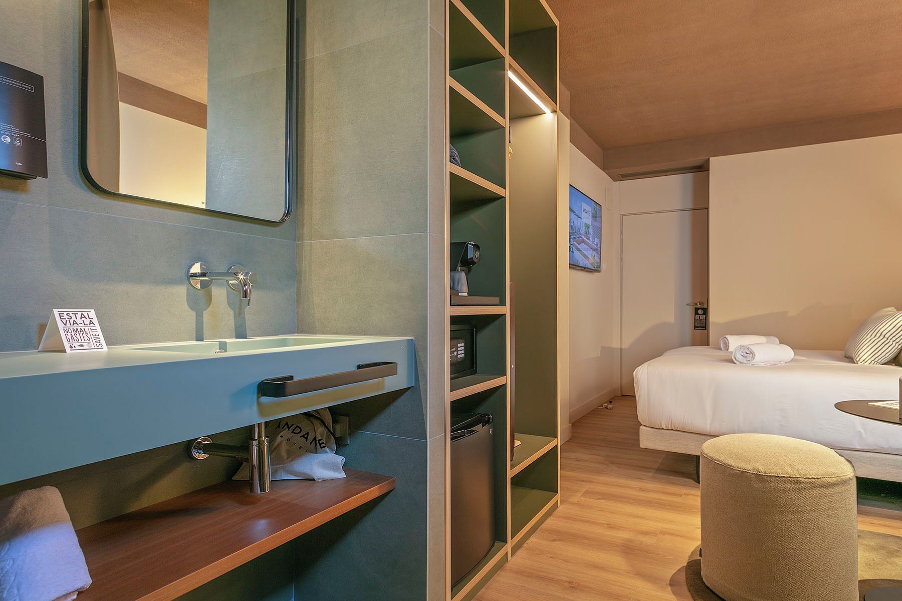 Habitación de hotel con baño, cama, mesita de noche y un pequeño otoman.