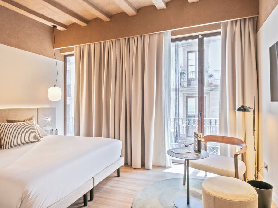 Habitación de hotel con cama, una mesita, una silla, un otoman redondo, una lámpara y vistas a la ciudad a través de amplios ventanales.
