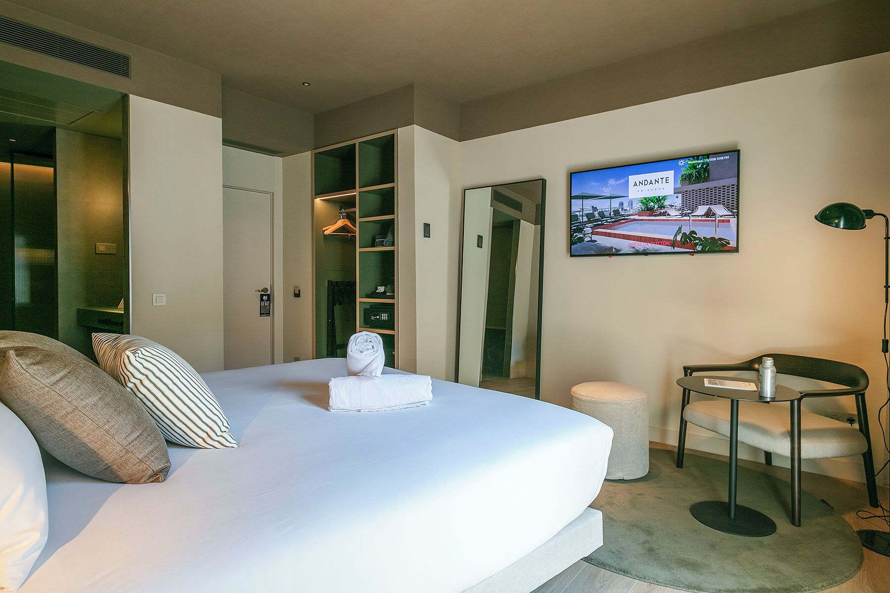 Quarto de hotel com cama, área de estar com uma cadeira, mesa pequena, televisão fixada na parede e armário com espelho.