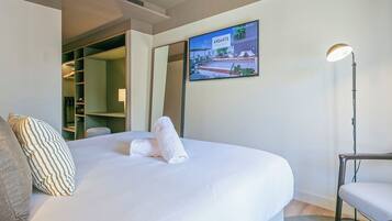 Habitación de hotel con cama, una silla, un escritorio, un espejo y un televisor que muestra un paisaje urbano.