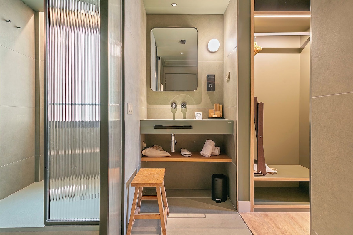 Un baño moderno con un espejo grande, un lavamanos, un taburete de madera y un cubo de basura.