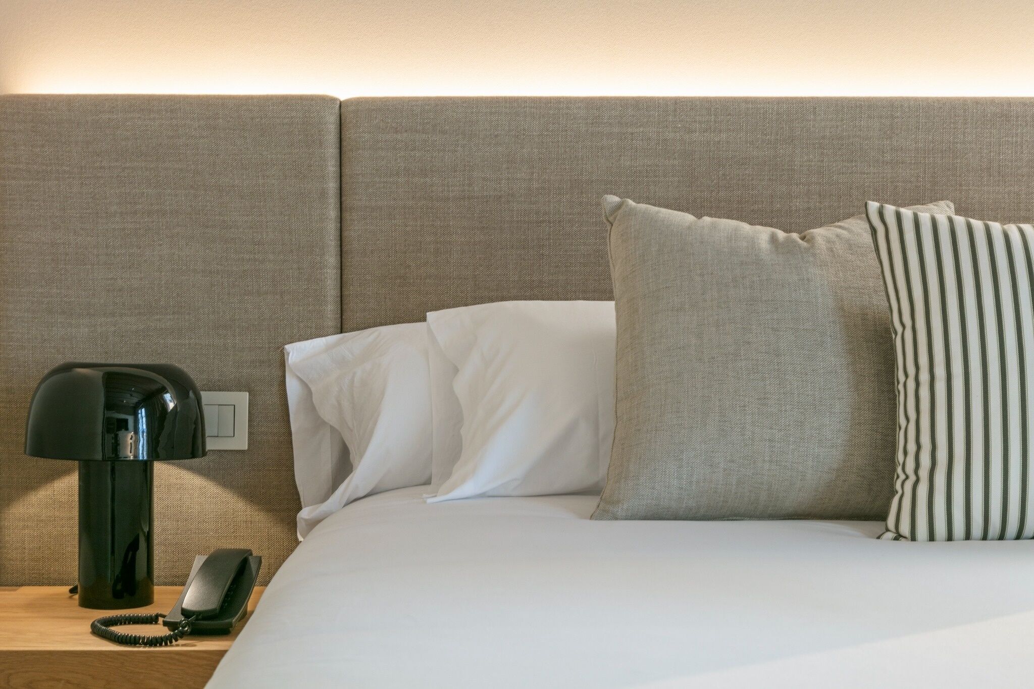 Habitación de hotel con cama, almohadas blancas y una a rayas, una lámpara y un teléfono en una mesita de noche.