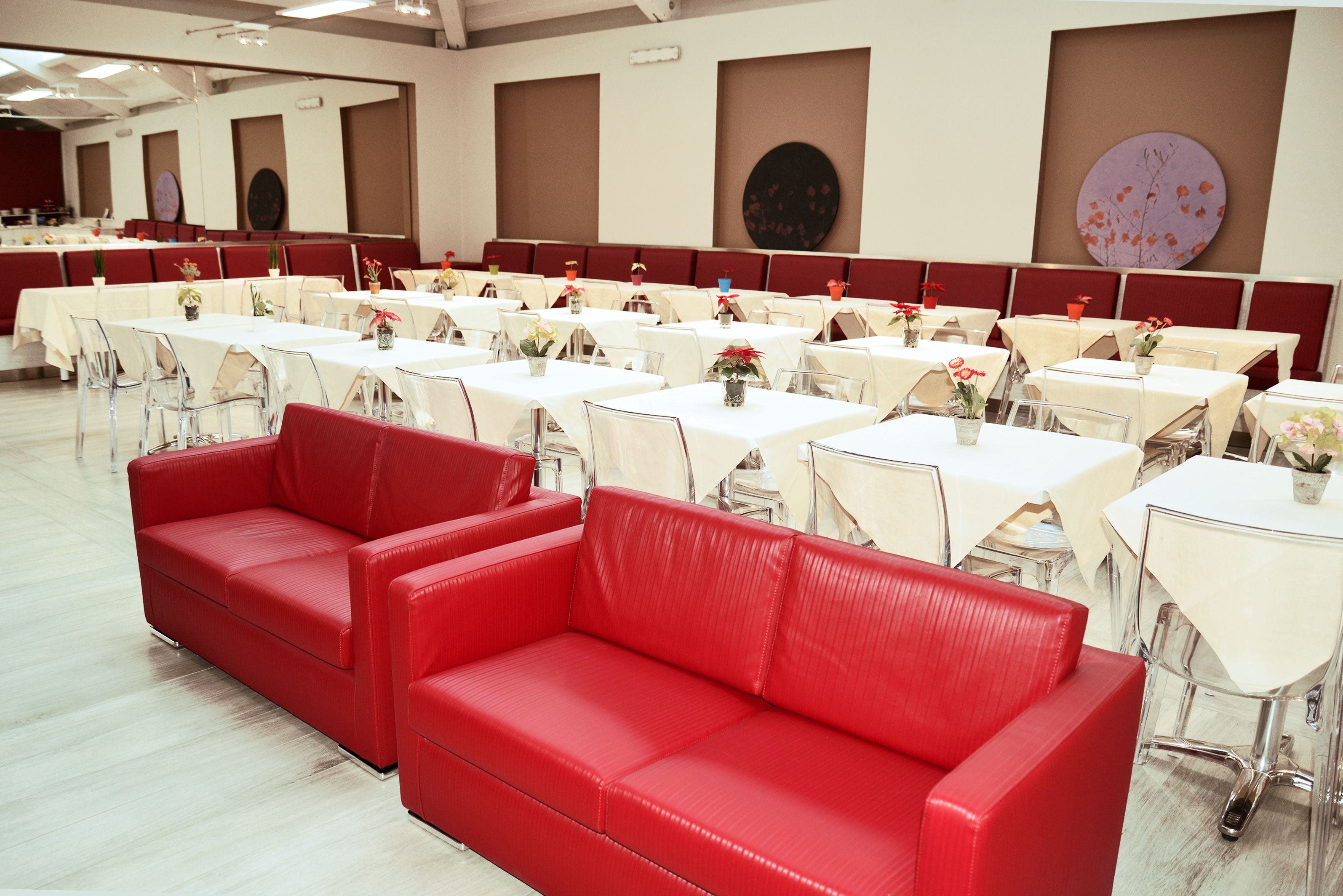 Ein geräumiger Festsaal mit Tischen und Stühlen, einem roten Sofa und dekorativen Wandbildern.