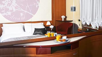 Quarto de hotel com cama, mesa de cabeceira com luminária, espelho, pequena mesa com itens de café da manhã e uma janela grande com cortinas.