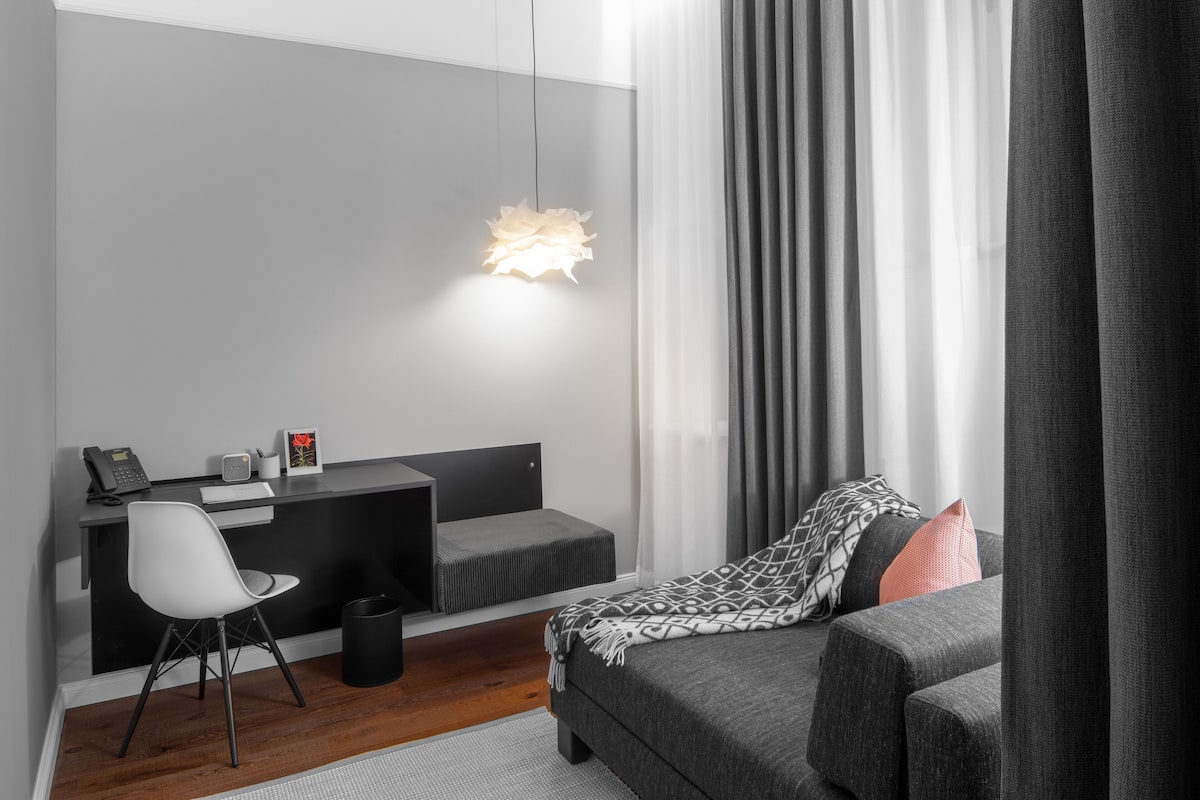 Un dormitorio moderno con un sofá gris, un escritorio negro con una silla, una lámpara colgante y una ventana con cortinas translúcidas.
