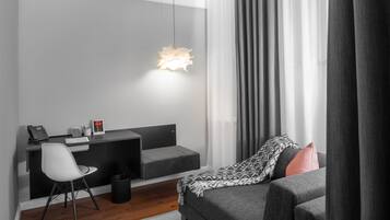 Une chambre moderne avec un canapé gris, un bureau noir avec une chaise, un luminaire suspendu et une fenêtre ornée de rideaux légers.