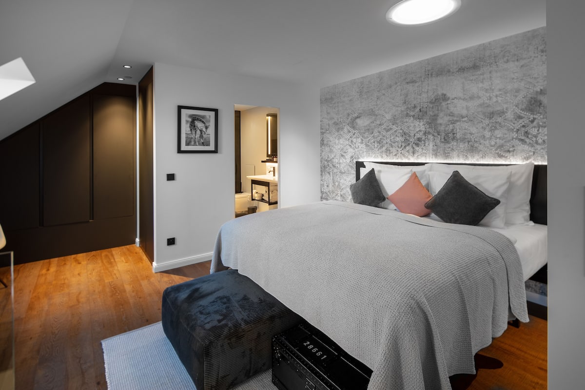 Un dormitorio moderno con una cama grande, cabecera con muchos almohadones, un armario oscuro y un tragaluz.