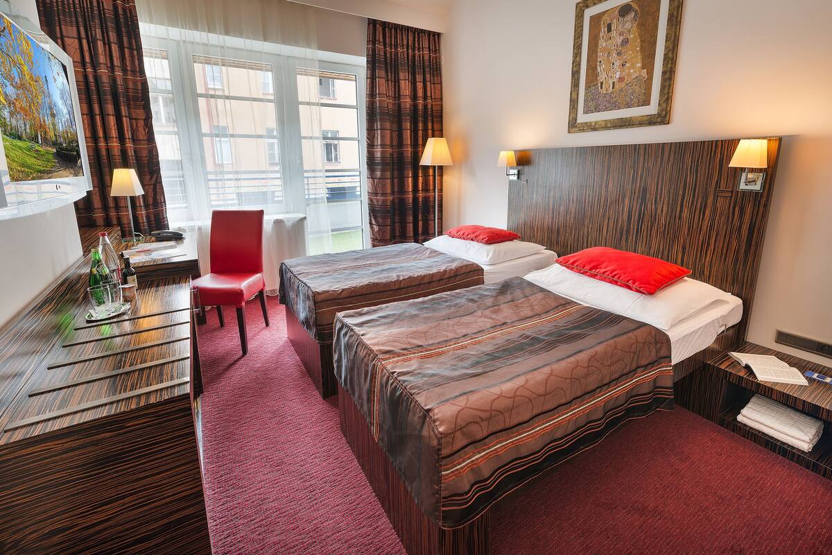 A hotel room with two beds, a desk with a chair, a window with curtains, and a painting on the wall.