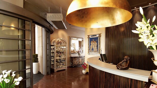 A reception area with a curved desk, a large gold pendant light, a glass display case, and a potted plant with white flowers. - EA Hotel Crystal Palace (Prague)