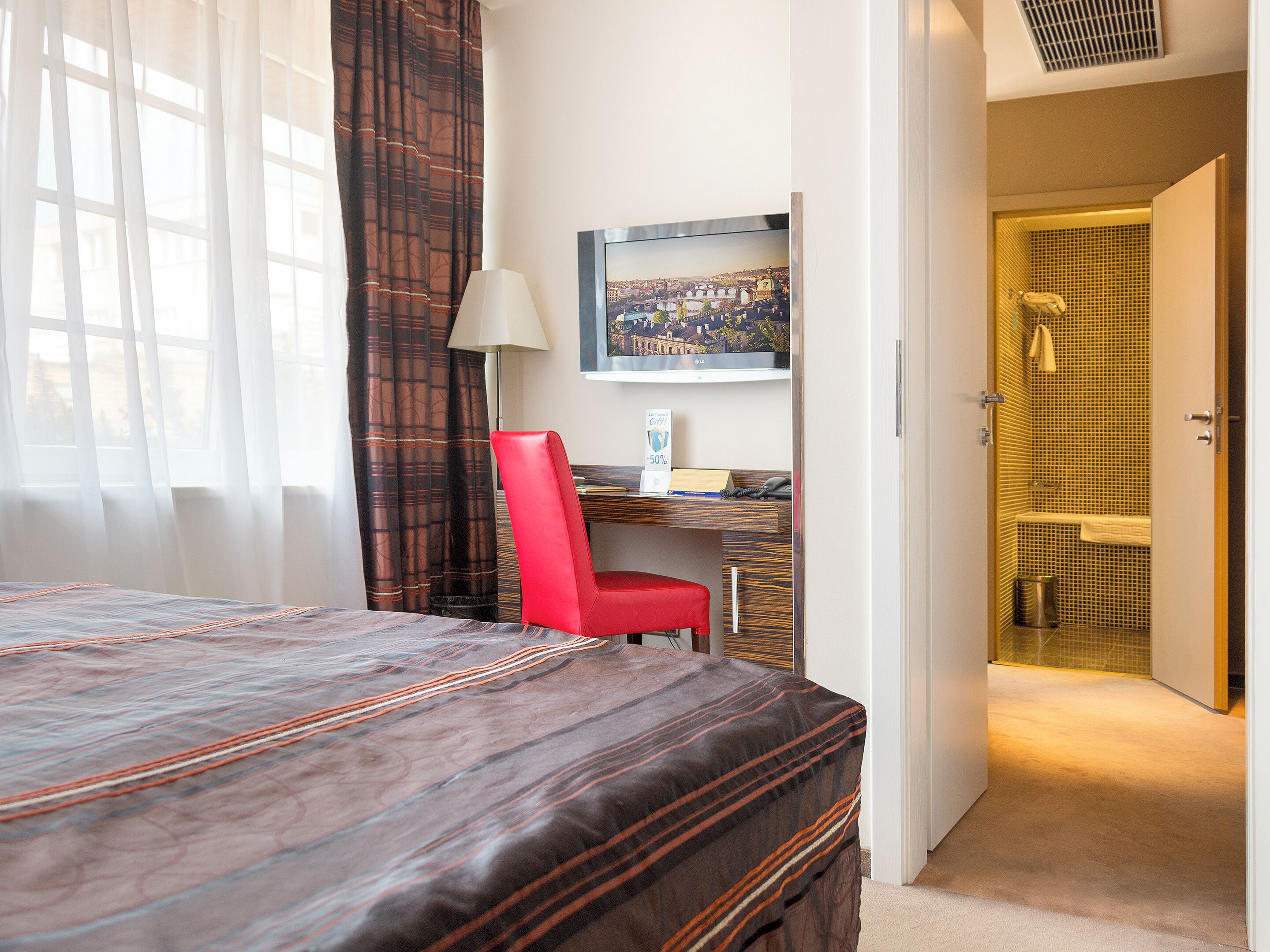 Una habitación de hotel con cama, escritorio, una silla roja, lámpara y baño visible a través de una puerta abierta.