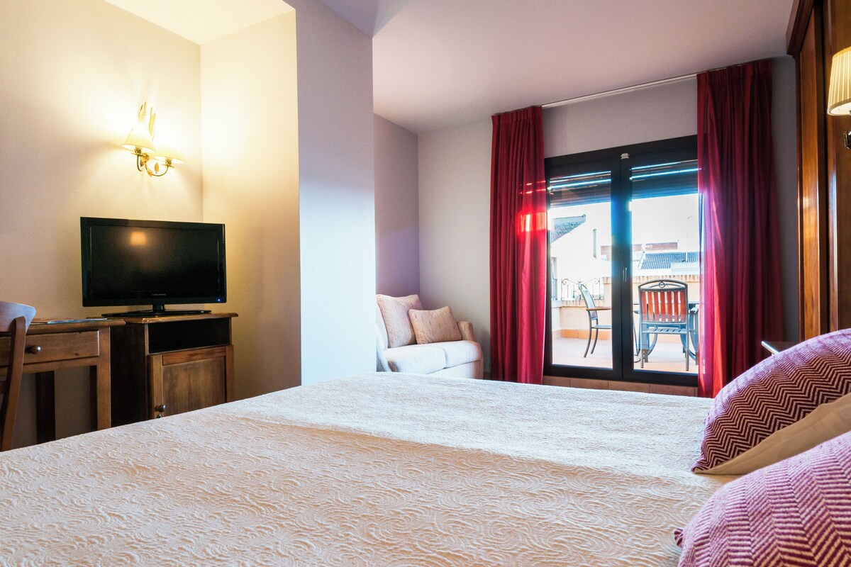 A hotel room with a bed, a desk with a television, a chair, and a view of a balcony with red curtains.