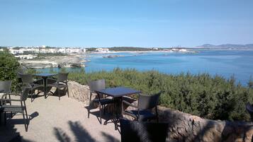 Un espacio al aire libre con sillas de mimbre y una mesa, con vista al mar y edificios a lo lejos.