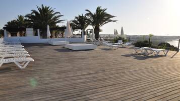 Una terraza de madera con sillones de descanso blancos y sombrillas, palmeras y vista del horizonte de la ciudad.