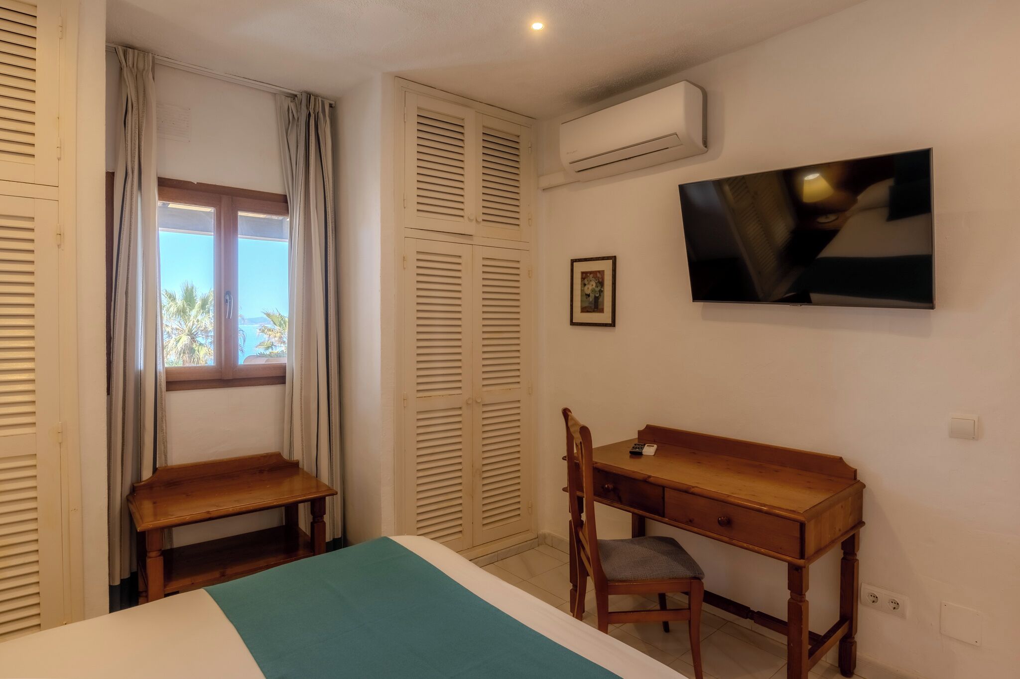 A bedroom with a bed, a desk, a chair, a window with curtains, an air conditioning unit, and a wall-mounted TV.