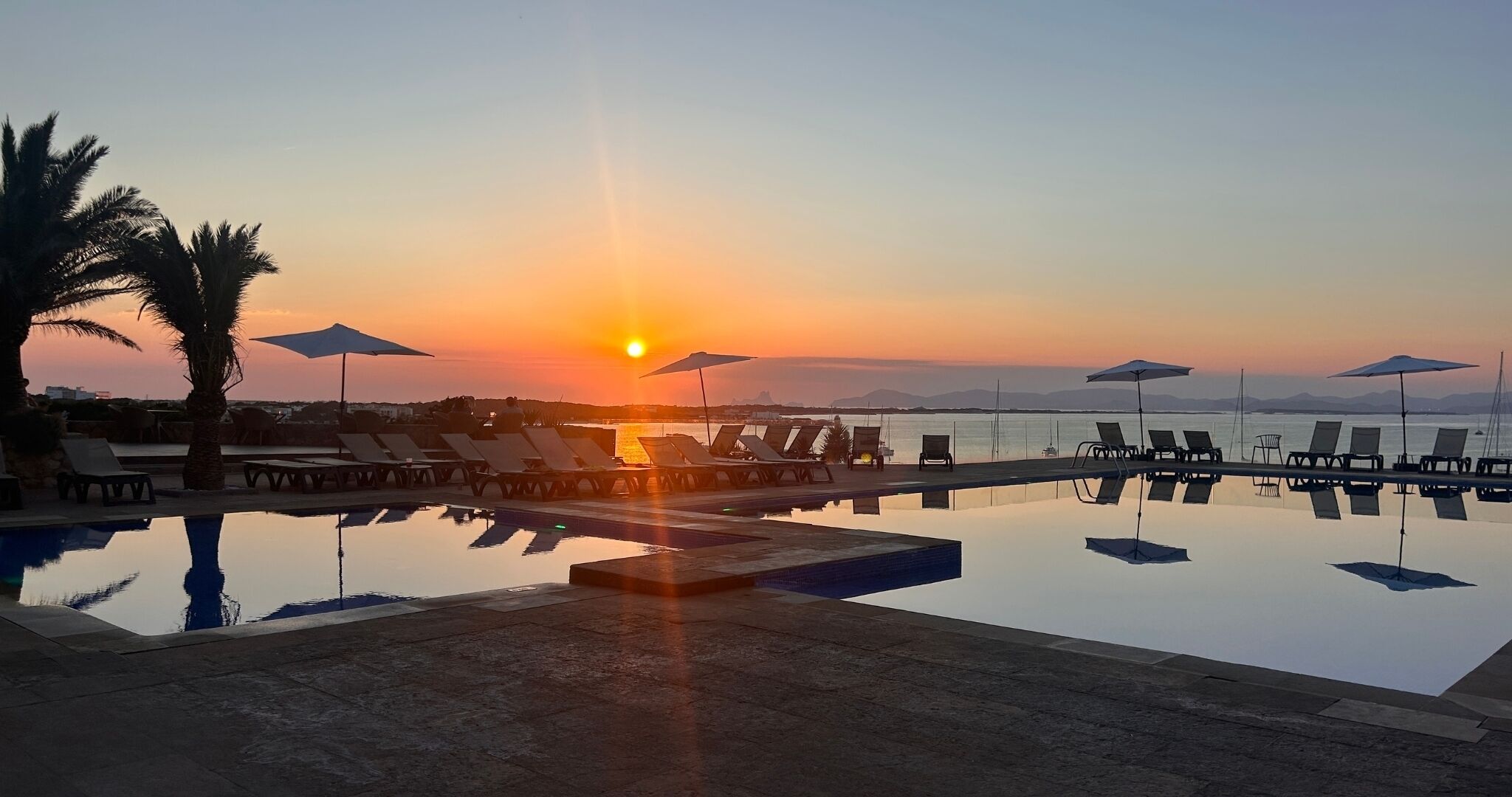 A poolside area with lounge chairs and umbrellas, overlooking a body of water at sunset.