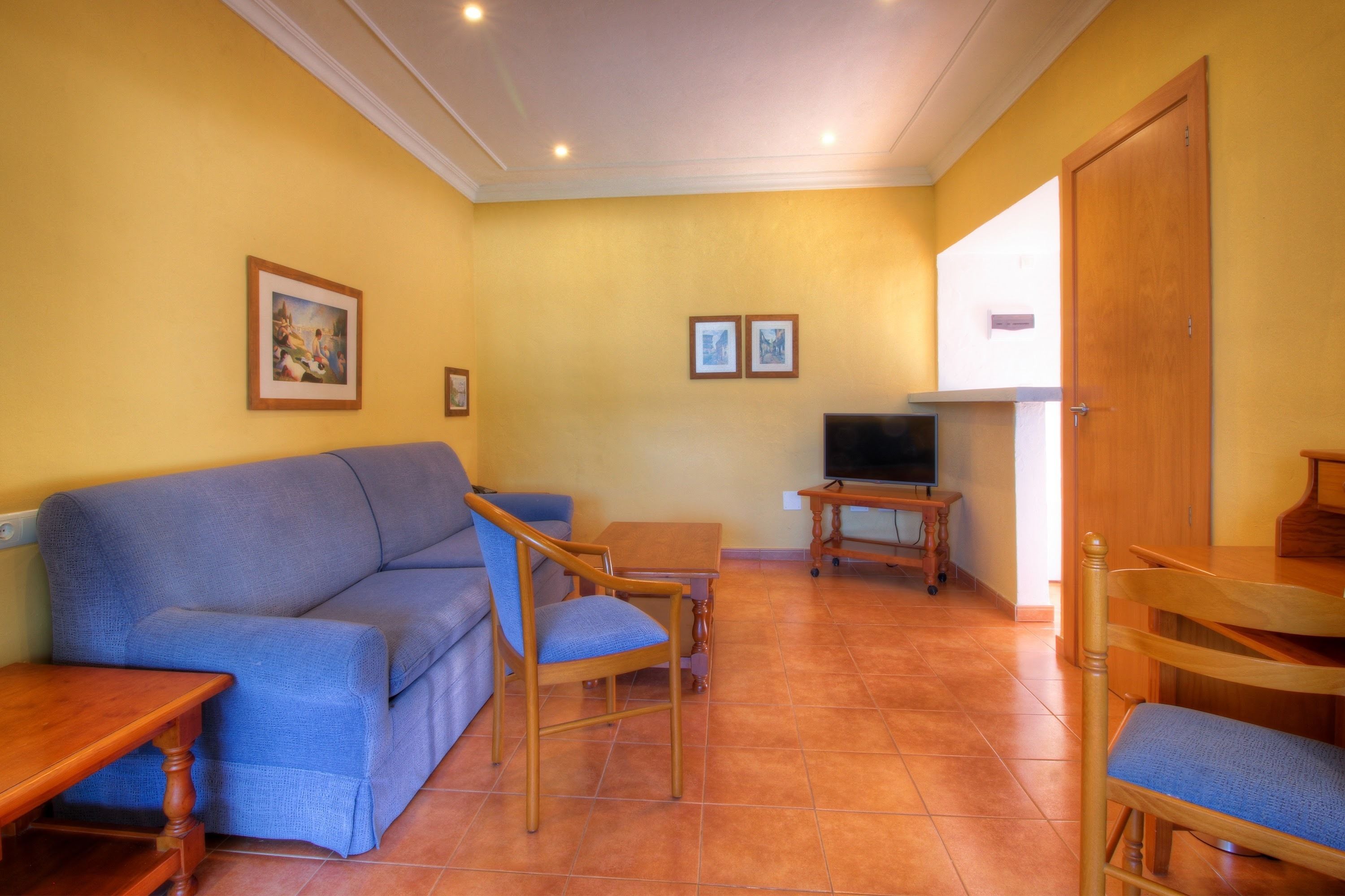 A living room with a blue sofa, wooden chairs, a TV, and two framed pictures on the wall.