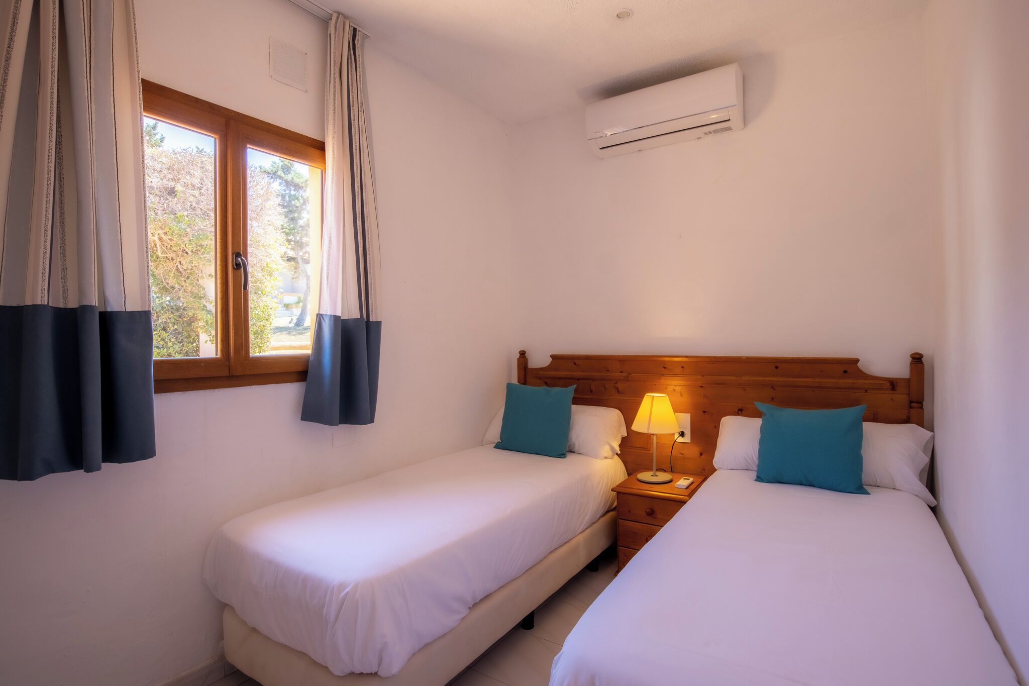 Ein Hotelzimmer mit zwei Einzelbetten, einem Holz-Kopfteil, einem Nachttisch mit Lampe und einer Wand-Klimaanlage.