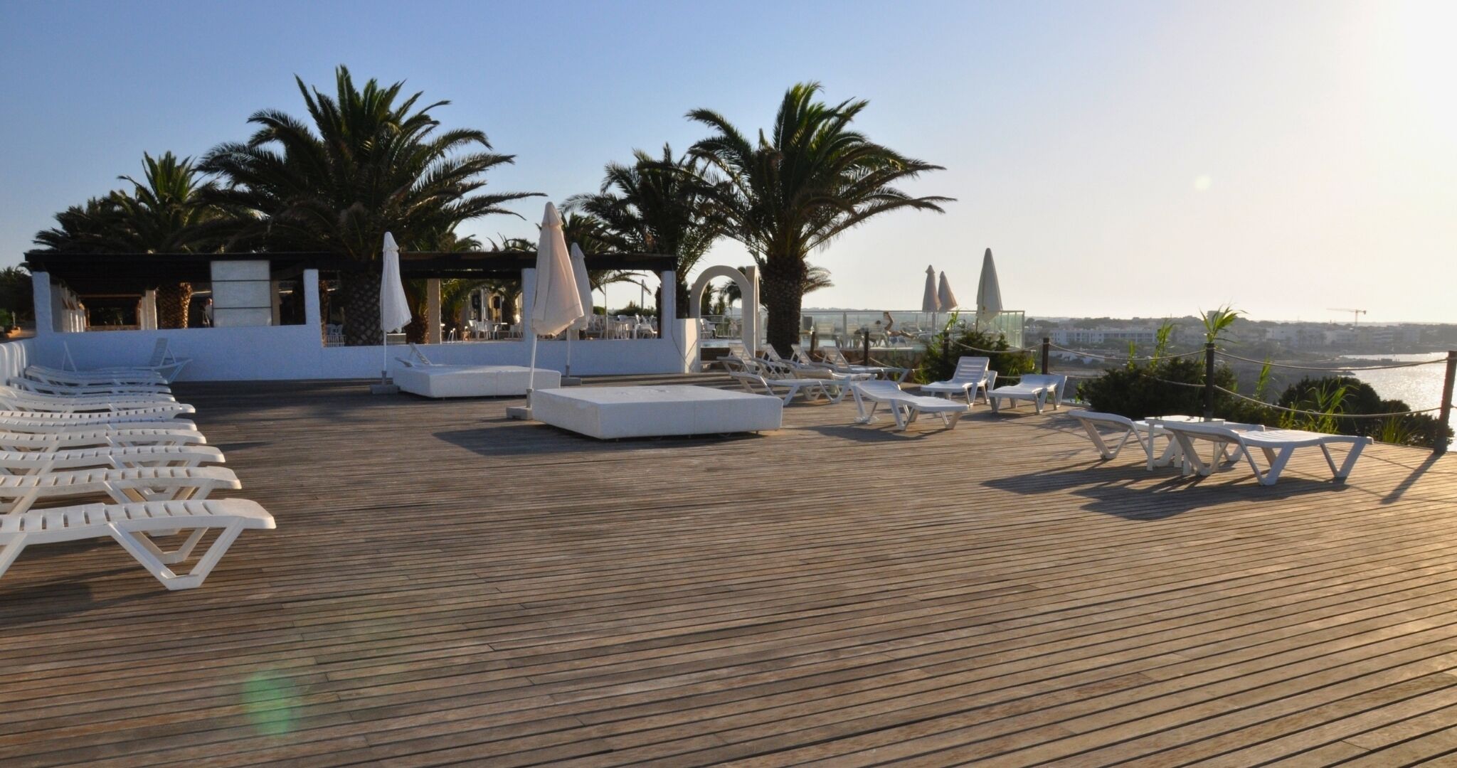Terraza de madera con sillas reclinables blancas y sombrillas, palmeras y vista a la ciudad.
