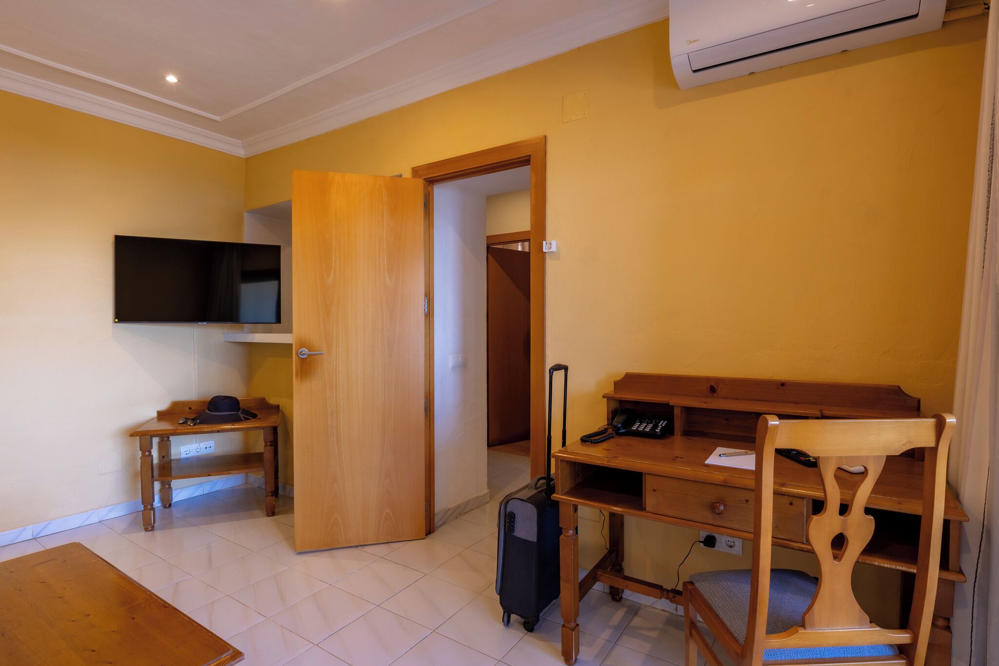 Una habitación de hotel con un escritorio de madera, una silla, un televisor, una maleta y un armario.