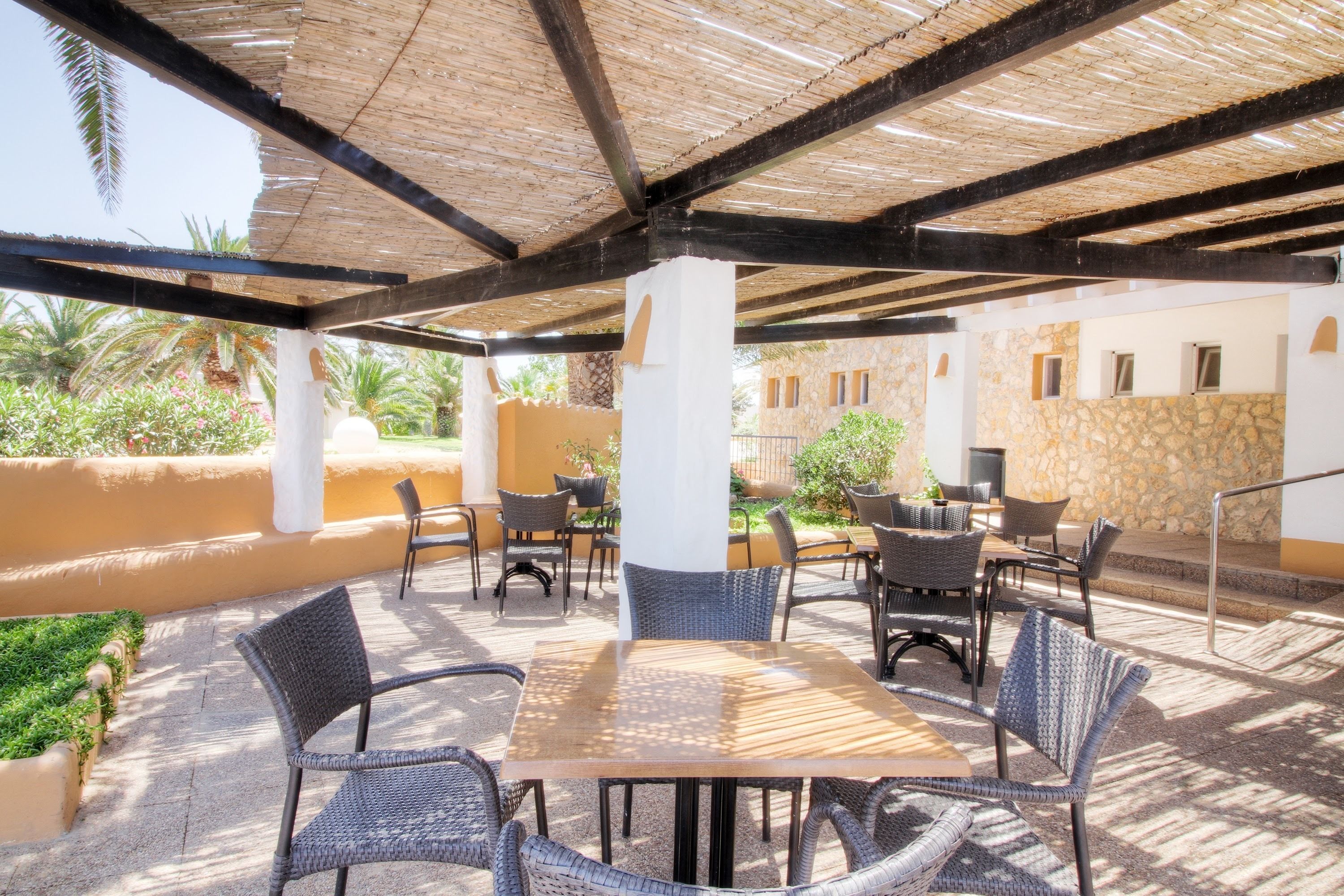 An outdoor patio with wicker furniture and a wooden table, surrounded by stone walls and palm trees.