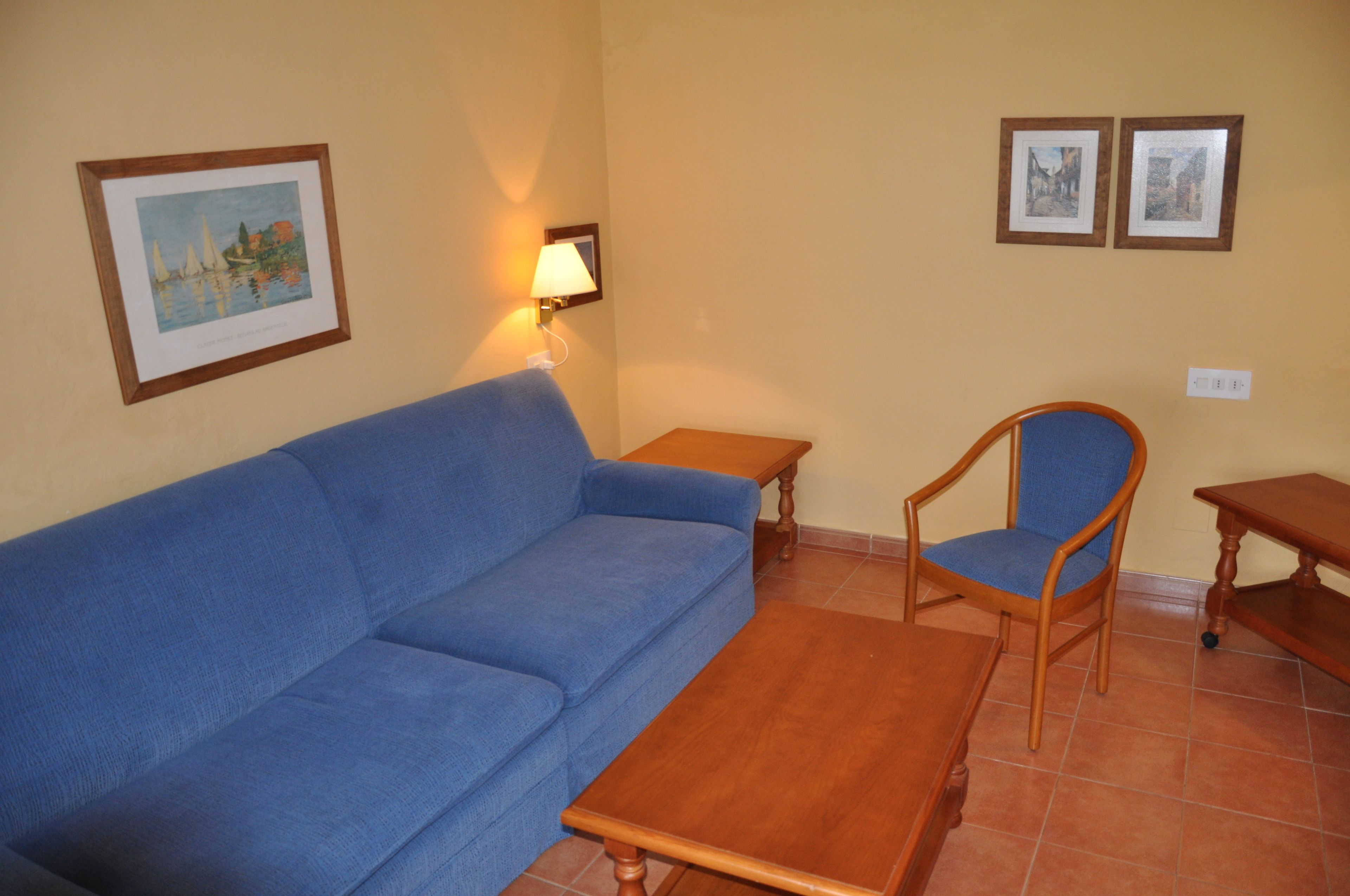 A living room with a blue sofa, wooden coffee table, and a wooden chair. There are two framed pictures on the wall and a wall-mounted lamp.