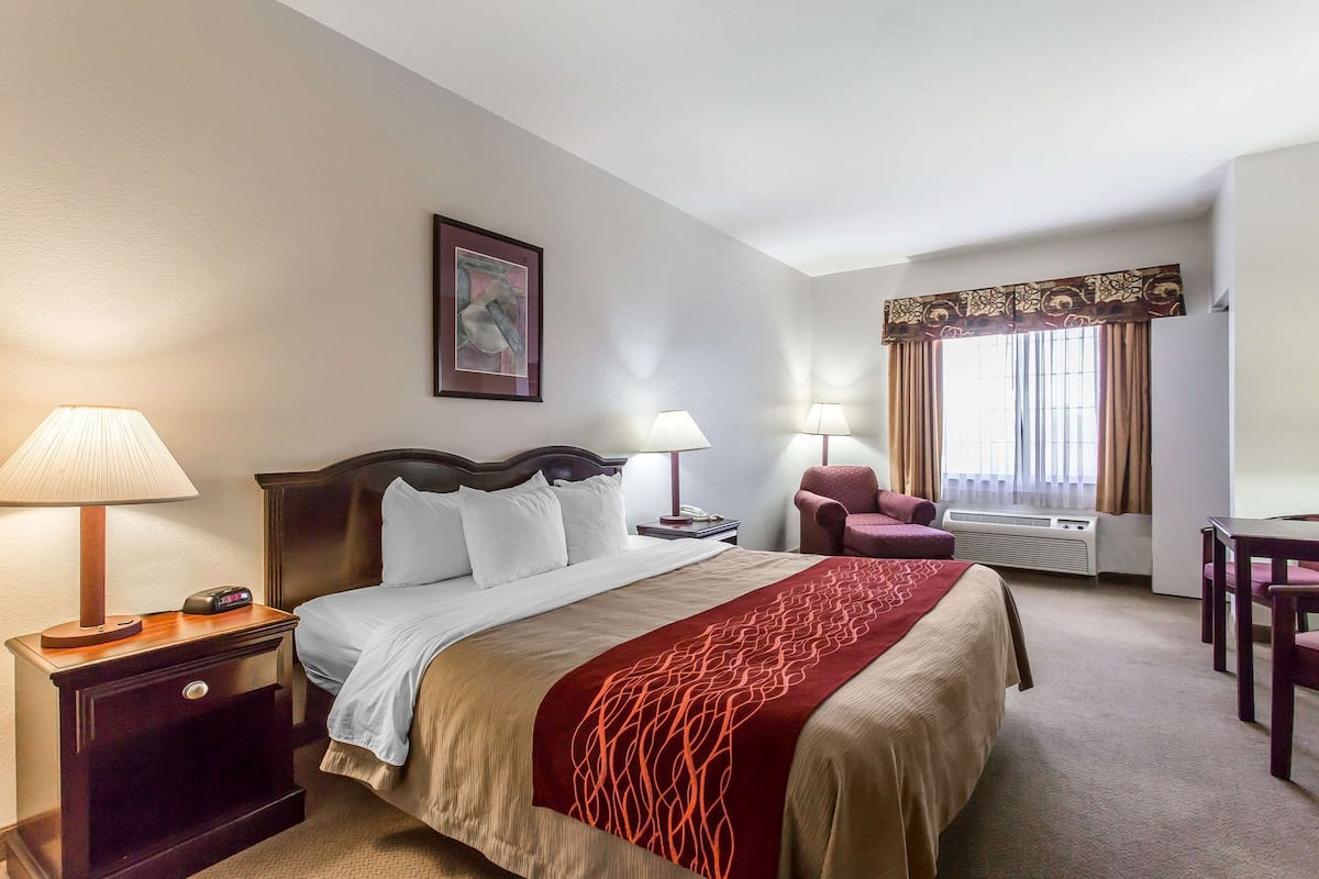 A hotel room with a bed, bedside lamps, a chair, a table, a picture on the wall, and a window with curtains.