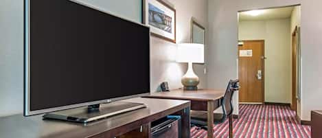A hotel room with a flat-screen TV, a desk with a lamp, a chair, and a patterned carpet.